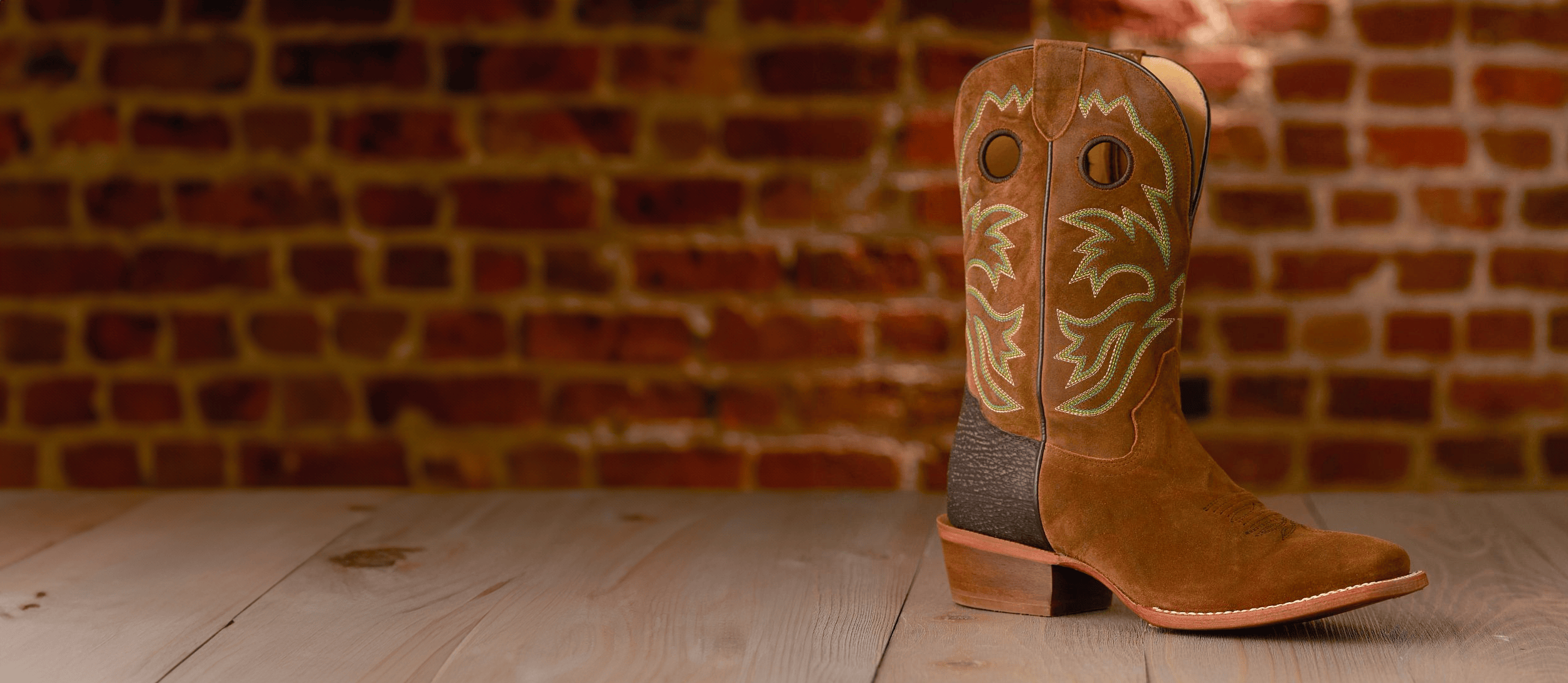 A single Hudson 11” Western boot in Crown in brown in front of a brick wall. Shop final sale. 